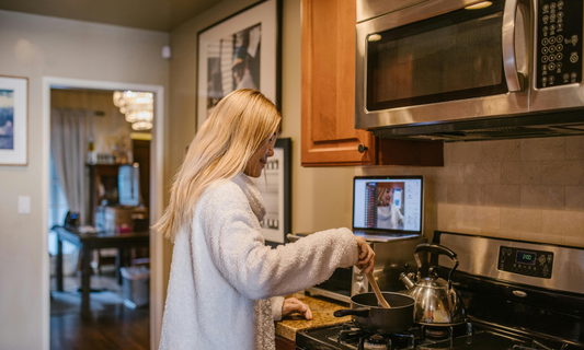 A woman in a white fleece jacket stirring a pot on a gas stove in a home kitchen while a laptop sits on the counter behind her