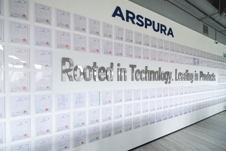 Arspura patent display wall with rows of framed certificates, ARSPURA lettering above, and the slogan “Rooted in Technology. Leading in Products.” in metallic letters.