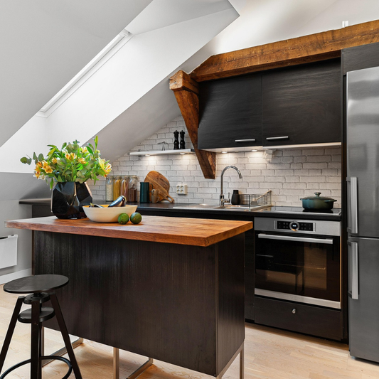 Small attic kitchen with sloped ceiling featuring dark wood cabinets, white subway tile backsplash, exposed wooden beams, under-cabinet lighting, wooden island with black base, and stainless steel appliances
