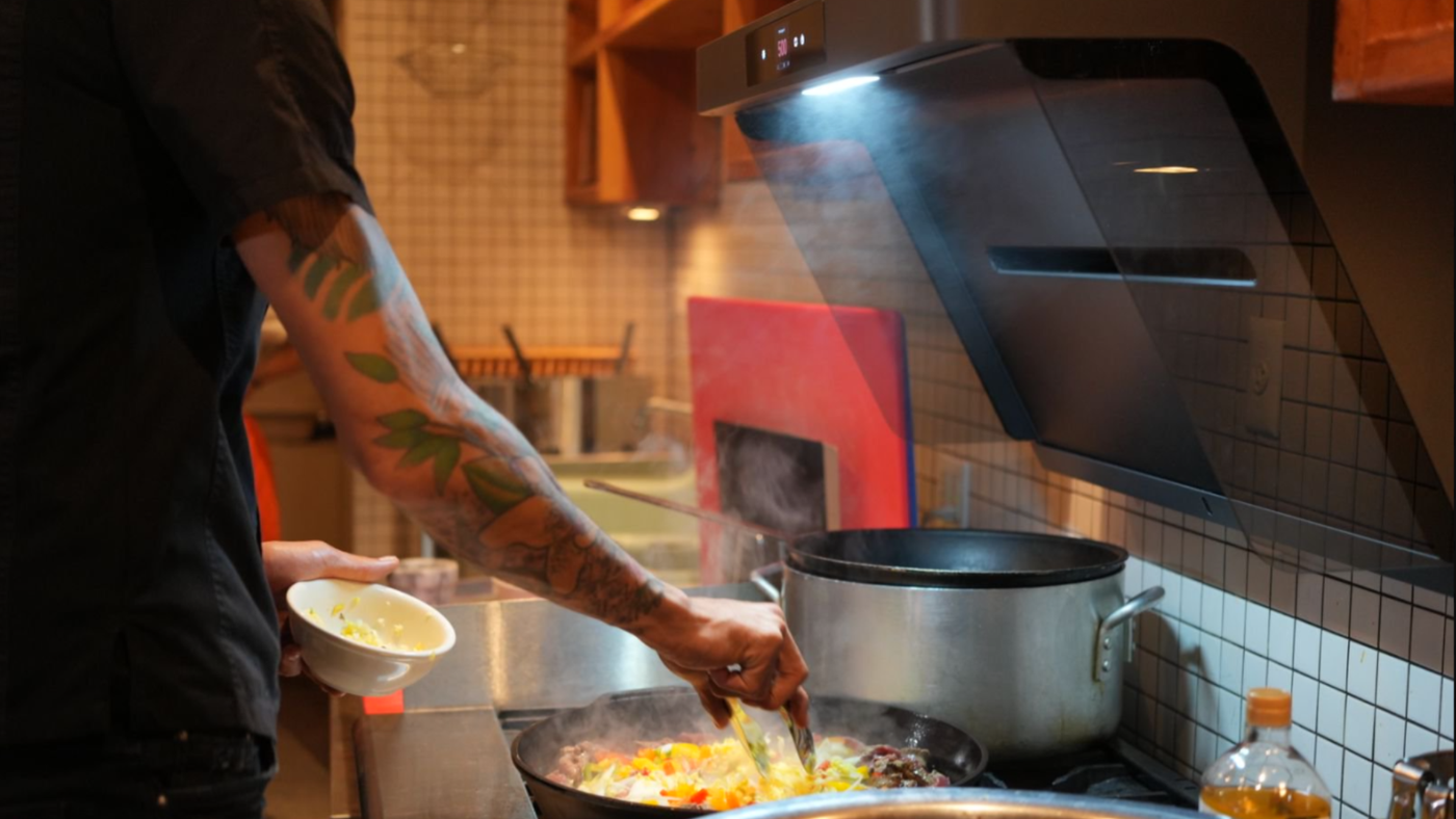 Close-up of a chef cooking under a range hood with visible steam and fumes