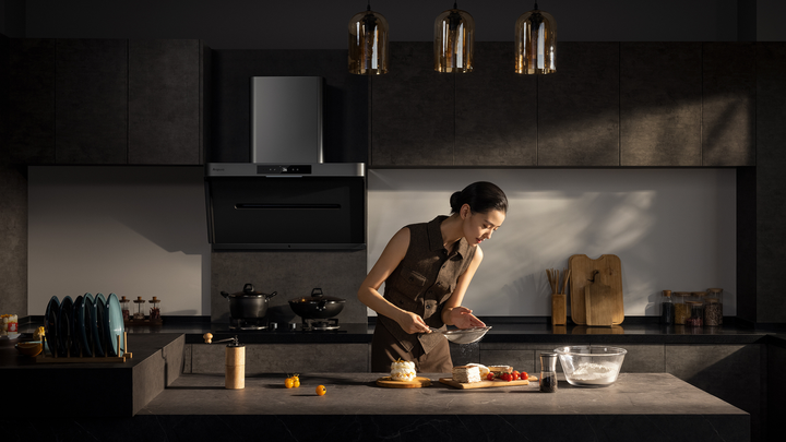 A woman in a brown vest sifting flour at a dark grey kitchen island in a sleek, modern kitchen equipped with a black Arspura range hood