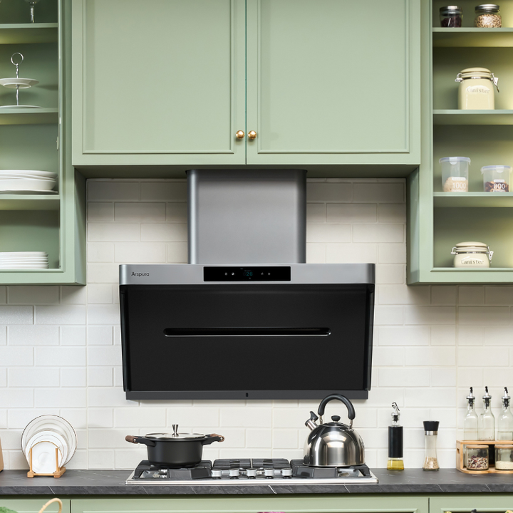Sage green kitchen cabinets with glass-front display shelves, black and stainless steel wall-mounted range hood above gas cooktop, white subway tile backsplash and dark countertop