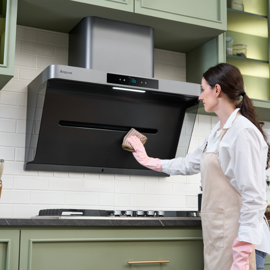 Woman in white shirt and apron using pink oven mitts to handle cookware on a gas stovetop beneath an Arspura wall-mounted range hood in a kitchen with sage green cabinets (duplicate angle)