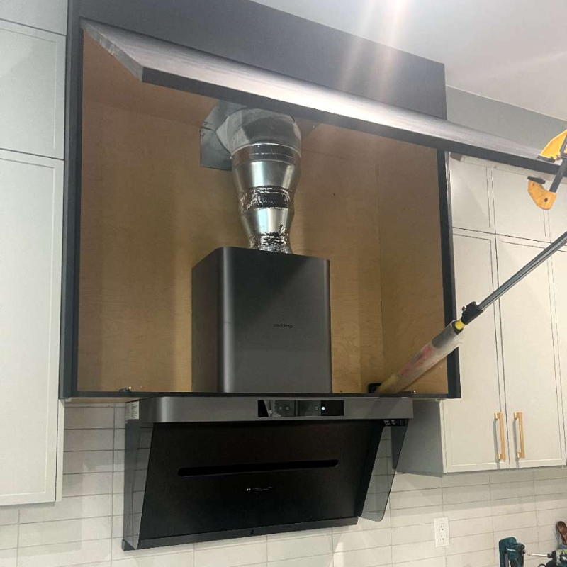 Black wall-mount range hood with rigid metal ductwork and 90-degree elbow installed above professional gas cooktop in white kitchen